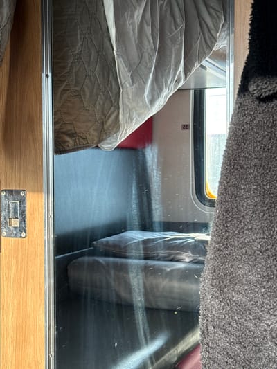 The image presents an interior view within a train sleeping compartment, located in Volkhov, Russia. A vertical, reflective glass panel dominates the center, bordered on the left by a light wood-grain panel to which a worn, silver-toned metal latch is affixed. The right side of the frame is partially obscured by a textured, dark grey, shaggy fabric.

Reflected within the glass, an upper bunk is partially visible, draped with a quilted, light brown blanket and a lighter, smoother sheet. Below this, two dark grey pillows are stacked on what appears to be a lower bunk or sleeping surface. A faint red object is also reflected behind the pillows. The number "32" is prominently displayed on an interior wall, visible through its reflection on the glass. To the right of the reflection, a vertical window with rounded corners, characteristic of railway carriages, admits bright daylight, indicating an external environment. The glass surface exhibits minor smudges, streaks, and particulate matter. No individuals are present in the scene.
