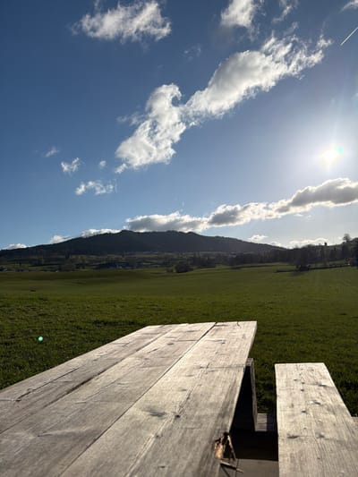 The image depicts an outdoor rural landscape in Gibloux, Switzerland, captured on a sunny day. In the foreground, a weathered wooden picnic table and an attached bench occupy the lower third of the frame, presented from a low-angle perspective. The wooden planks exhibit visible grain, cracks, and discoloration. Extending from the mid-ground to the background is an expansive, undulating green grassy field, illuminated by direct sunlight. In the distance, a prominent dark green, tree-covered mountain range forms the horizon, with several small structures or buildings discernible at its base. The sky above is clear blue with scattered white cumulus clouds. A bright sun is visible in the upper right quadrant, producing a strong lens flare and emanating light across the scene. A faint white contrail from an aircraft is present in the upper right sky. No individuals or animals are present, and no specific actions or interactions are occurring.