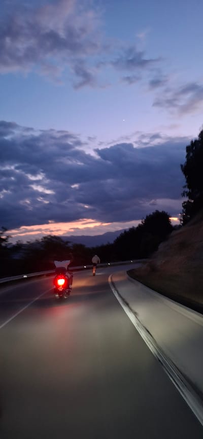 At dusk, a person rides a motorcycle on a paved road that curves to the right. The motorcycle's taillight is illuminated. Another person is visible further up the road, walking in the same direction as the motorcycle. The sky is a gradient of deep blue and purple, with scattered clouds and a sliver of the moon visible. Hills and trees are silhouetted against the horizon. The road has white lane markings and reflectors. The scene appears to be on the outskirts of a city, possibly Valkosel, Bulgaria, given the mountain and wooded landscape.