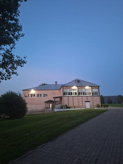 The image depicts an exterior view of a building at dusk in Атамановка, Russia. The building, with a light pink facade and a grey metal roof, features multiple windows and an entrance with an awning. The upper section of the building has large, multi-pane windows, suggesting a large interior space. The ground in front of the building is a manicured lawn that slopes gently downward, with a paved walkway to the right. The sky is a clear, deep blue, transitioning to a lighter shade near the horizon, indicating the twilight hours. A large tree with dark foliage partially obscures the upper left corner of the frame. The scene is devoid of any visible people or animals.