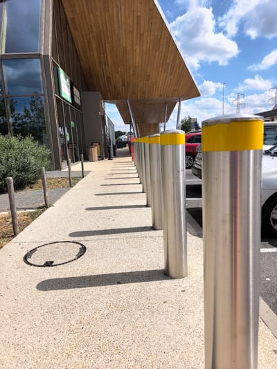 The image shows a line of bollards with yellow tops along a sidewalk in front of a modern building with a wooden canopy. The bollards are made of brushed metal and are spaced evenly apart, creating a visual barrier between the sidewalk and a parking lot. In the background, there are cars parked in designated spots, and beyond that, there are power lines and trees. The sky is blue with fluffy white clouds. The location is identified as Briis-sous-Forges, France. The scene appears to be an outdoor public space, possibly a commercial area or a community center, with a focus on pedestrian and vehicular access control. There are no people or animals visible in the image, and no actions or interactions are taking place. The environment is clean and well-maintained, with clear shadows cast by the bollards and the building's canopy, indicating a sunny day.