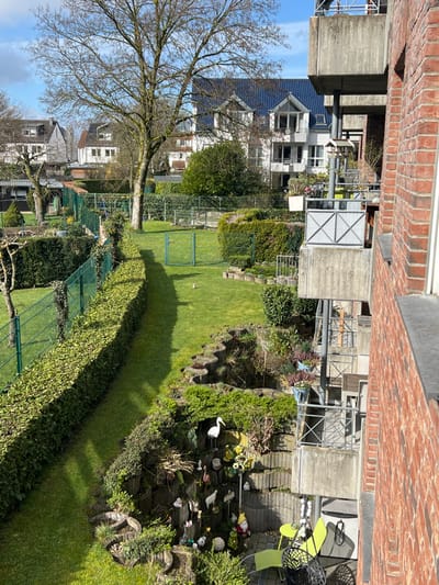 The image depicts a residential outdoor scene in Würselen, Germany, observed from an elevated vantage point, likely a balcony or upper window. The foreground on the right consists of a red brick building facade with a concrete balcony structure featuring metal railings. A lower balcony on the right side contains a pale green table and two matching chairs, along with a hanging black net object. Multiple concrete balconies are visible, some equipped with bird feeders and potted plants.
The midground features a series of meticulously maintained gardens under bright sunlight. A long, trimmed green hedge runs diagonally from the bottom left to the upper center-left, bordered by a green wire fence. Adjacent to this fence is a narrow grassy path. A large, bare deciduous tree stands centrally in the mid-distance. The gardens contain expansive green lawns, various shrubs, and garden ornaments, including a prominent white stork statue, multiple garden gnomes, flamingos, ducks, and mushrooms, positioned among tiered concrete planters filled with vegetation. A smaller green wire gate is visible within a fenced area on the left side of the lawn.
In the background, several multi-story residential buildings are visible, characterized by white facades and dark-colored roofs, some featuring solar panels. These buildings are interspersed with other trees, both deciduous and evergreen. No individuals are present in the depicted scene.