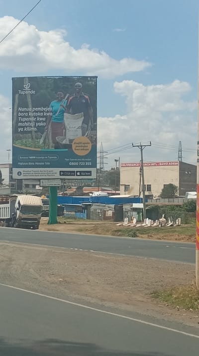 The image shows a "Tupande" billboard, advertising agricultural inputs. A man and woman are depicted carrying farm supplies. The billboard includes text in Swahili promoting buying better farm inputs through Tupande. It also mentions opportunities to receive credit for inputs and a toll-free number. A truck passes on the road below. A building labeled "DELIVERANCE CURCH INTL - BANGLADESH" is visible in the background, along with overhead power lines.