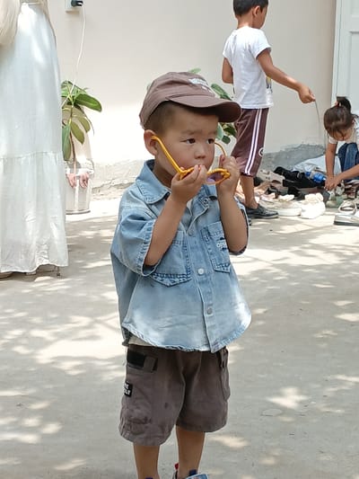 This image, taken in Osh, Kyrgyzstan, depicts a young boy wearing a brown baseball cap and a denim shirt. He is holding a pair of yellow sunglasses up to his face, seemingly examining them. The boy is also wearing brown shorts. In the background, another child, a boy, is wearing a white t-shirt and dark shorts with white stripes, and is interacting with a pile of shoes and bags. Further in the background, a young girl in a white shirt and blue jeans is also engaged with the shoes. A person in a long white dress stands to the far left of the frame. The ground is a concrete surface with dappled sunlight and shadows from overhead foliage. The overall setting appears to be an outdoor area, possibly a yard or courtyard, with a wall in the background.