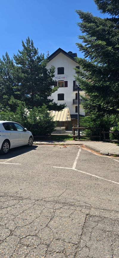 In Sallent de Gállego, Spain, a white hatchback car is parked in a cracked asphalt parking lot. Behind the car, a white building with a dark roof, identified by the signage "EDIFICIO ARIES", stands partially obscured by tall green pine trees. The building features multiple windows with dark shutters and a small balcony. In front of the building, a light-colored, angled canopy covers an entrance area. A lamppost is positioned near the building's entrance. The scene is set under a clear blue sky on a sunny day.