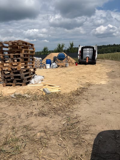 This is a ground-level, outdoor, medium shot of a construction site under a cloudy sky. In the foreground, dry grass and dirt make up the ground. To the left, a large stack of wooden pallets is visible. In the mid-ground, there are piles of sand and cement bags, a blue cement mixer, and a white van with its rear doors open, containing a large transparent water tank with a red pipe extending from it. A red canister also sits near the sand pile. In the background, trees and a field of crops are visible, with a dark fence running along the tree line. The scene is in Opatów, Poland.