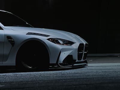 This media file captures a low-angle, close-up view of the front-right quarter of a modern light grey/white BMW M4 car. The vehicle is stationary and positioned against an extremely dark, almost black, background, suggesting either a night setting or a dimly lit indoor environment in Karadjovo, Bulgaria.
The car's design is prominent, featuring a distinctive LED headlight with an amber/orange accent and a bold, vertical kidney grille. The front bumper incorporates aggressive styling with complex lines, visible air intakes, and a black front lip spoiler that extends along the bottom edge. On the front fender, a black, multi-slatted vent is visible, complemented by a longer, angular vent integrated into the hood, both contributing to the vehicle's performance-oriented aesthetic. The front wheel, partially visible, is entirely black, creating a stark contrast with the light-colored bodywork.
The ground beneath the car is a textured surface, possibly asphalt or concrete, appearing coarse and somewhat worn, with subtle variations in tone that could indicate tire marks or dampness. The lighting is focused on the car, highlighting its contours and details while leaving the background in deep shadow, emphasizing the vehicle's form. No people are visible, and there is no discernible text present in the image. The overall impression is one of sleekness, power, and high-performance automotive design.