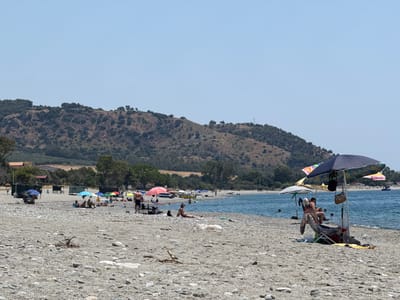 The image depicts a sunny day at a pebble beach in Bova Marina, Italy. In the foreground, a beach chair is occupied by two individuals under a large umbrella. Scattered across the beach are various beach umbrellas, some with people beneath them, and individual sunbathers. The sea is visible in the background, with a few people swimming or wading. Beyond the beach, a densely wooded hillside rises, with some buildings and structures nestled amongst the trees. The sky is clear and blue.
