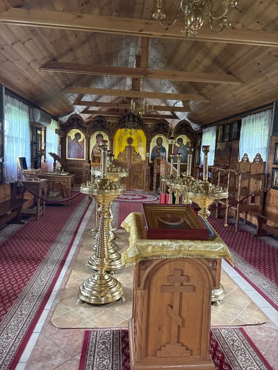 The image shows the interior of a wooden church in Rybaki, Poland. The church features traditional Orthodox Christian iconography and furnishings. A large iconostasis, or icon screen, made of carved wood and adorned with religious paintings, dominates the background. Several tall, ornate brass candelabra stand in the foreground, some with candles. In the center of the foreground, a wooden lectern with an Orthodox cross carved into its surface holds a religious book covered with a gold cloth. Red carpet with intricate patterns covers the floor in aisles, with tiled flooring visible in other areas. Wooden benches and chairs are positioned along the side walls, and white curtains hang in front of windows. The ceiling is constructed of wooden planks. There are no people visible in the image.