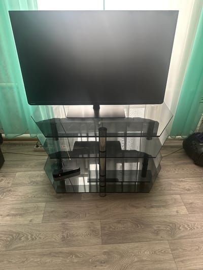 A television is centered on a three-tiered glass TV stand, with a black base. The TV stand is positioned on a light brown wooden floor, against a backdrop of light teal and white curtains. The lower shelf of the stand holds a black electronic device with a red indicator light and a remote control. To the right of the TV stand, a dark grey vacuum cleaner is visible. The lighting appears to be natural daylight, coming from the window behind the curtains. The location is described as Yazykova Pyatina, Russia.