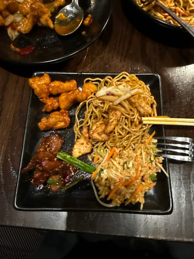 The image shows a meal served on a square black plate. The meal consists of three distinct components: noodles mixed with pieces of cooked chicken, fried rice containing carrots and green onions, and pieces of glazed chicken and beef. Utensils, including chopsticks and a fork, are visible atop the plate. Partially visible in the image are additional black dishes containing similar food items, set on a dark wooden surface. The location is the city of Chula Vista, United States of America.