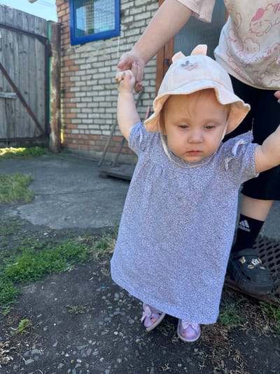 An outdoor, eye-level, medium shot shows a toddler and an adult in a yard. The toddler, wearing a light pink sun hat with rabbit ears and a gray speckled dress, is being supported by the adult's hands as they stand on a concrete pathway. The adult, partially visible from the chest down, is wearing black leggings and a white t-shirt with a graphic print of cartoon cats. The toddler's shoes are light purple with pink bows. The background features a brick building with a blue-framed window, a wooden fence, and overgrown grass. The location is identified as Novoyeniseyka, Russia.