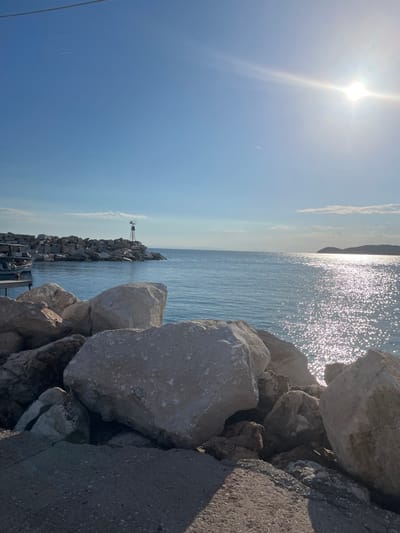 The image depicts a rocky coastline with the sea and sky dominating the frame. In the foreground, large, weathered rocks are piled up, with some smaller stones and a paved surface visible. A body of water, likely the sea, stretches out to the horizon. The water is calm with sunlight reflecting off its surface, creating a shimmering effect. In the middle ground, a man-made structure, possibly a breakwater or pier, is constructed from large rocks. A small lighthouse or navigational marker stands atop this structure. A boat is moored to a wooden dock on the left side of the image. The sky is a clear blue with a bright sun shining in the upper right corner. A faint contrail from an aircraft is visible in the upper left portion of the sky. The scene appears to be in Ποτός, Greece, a coastal town known for its picturesque beaches and clear waters.