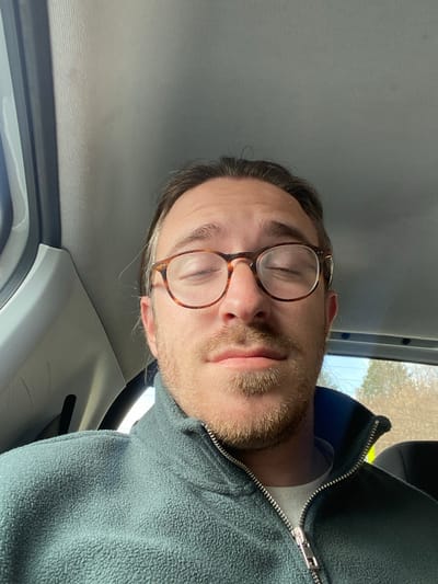 A Caucasian male, estimated to be in his late 20s to early 40s, is seated inside a vehicle in Kolašin, Montenegro. He has dark brown hair with visible graying at the temples, a light brown beard and mustache, and wears tortoise-shell rimmed glasses. His eyes are closed. He is dressed in a teal-green fleece jacket with a silver zipper, worn over a light-colored undershirt. The vehicle's grey headliner is visible overhead, and a blurred exterior scene featuring trees and a blue sky is visible through a rear window on the right side of the image. The scene is illuminated by natural light.