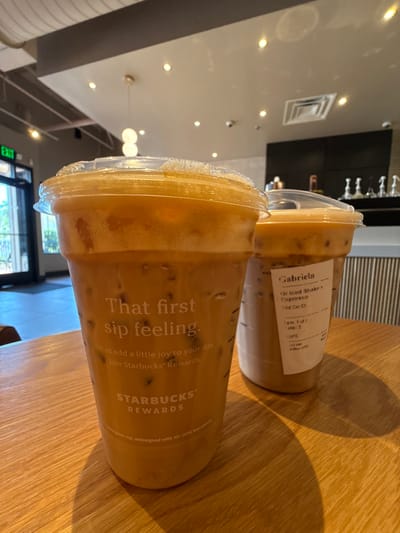 The image displays two Starbucks iced beverages on a light-colored wooden table inside a Starbucks coffee shop located at 1302 Trade Center Ct, Naples, FL 34109, United States.
In the foreground, a large, clear plastic Starbucks cup with a dome lid contains a light brown, frothy iced beverage with visible ice cubes. The cup features the printed text "That first sip feeling." and "STARBUCKS® REWARDS." Behind it, a smaller, clear plastic Starbucks cup with a flat lid also holds a light brown iced beverage with ice. This cup has a white label wrapped around it, identifying the order for "Gabriela" as a "Gr Iced Shake I Espresso," prepared at "03:15 PM" on "4 Mar 2020" at the "Trd Car Ct" location.
The background shows the interior of the coffee shop, including a glass exit door with an illuminated green "EXIT" sign to the left. The ceiling features recessed lighting and exposed light gray ductwork. A counter area with dark surfaces and silver implements is visible in the distant background, alongside a white, vertically ribbed wall section. No individuals are present in the scene.