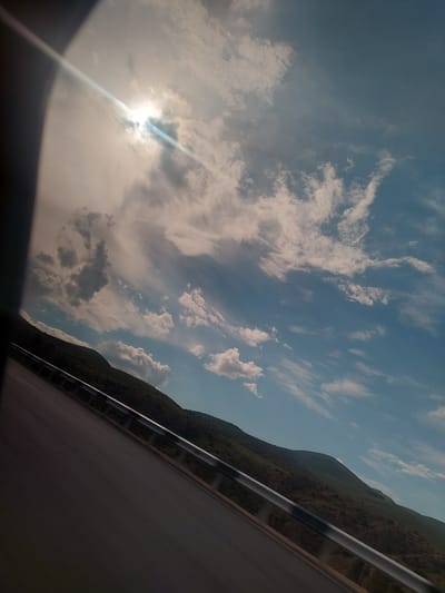 This dynamic photograph, likely taken from a moving vehicle, captures a scenic mountainous landscape near Yelpin, Armenia, under a bright, partly cloudy sky. The image is tilted counter-clockwise, creating a diagonal composition.
The upper left half of the frame is filled with a vast expanse of sky, featuring a mix of blue patches and numerous white and grey clouds of varying densities and shapes. The sun, partially hidden behind clouds in the far upper left corner, emits a powerful glare and a distinct, long lens flare that streaks diagonally across the sky.
Along the right side of the image, a dark green mountain range stretches into the distance, its textured slopes indicating rugged terrain and vegetation. In the foreground, occupying the lower right, a grey, paved road or highway is visible, bordered by a metallic guardrail consisting of two horizontal bars. The blur along the bottom left edge suggests the interior of the vehicle from which the photo was captured.
The strong sunlight indicates daytime, likely late morning or afternoon. The weather appears fair, with scattered clouds. The scene conveys a sense of travel through a picturesque, natural environment. No visible text is present.