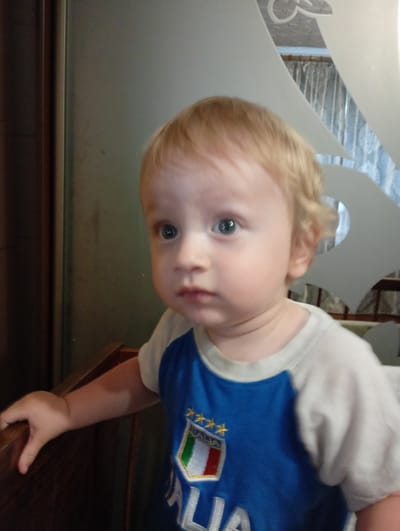 A close-up, medium shot captures a young Caucasian toddler with fair skin and blonde hair, looking to the right of the frame. The toddler is wearing a blue and white raglan t-shirt with an Italian flag emblem on the chest, featuring green, white, and red stripes with three gold stars. The toddler's left hand is resting on a dark brown wooden surface, possibly a chair or table edge, which occupies the bottom left portion of the image. The background is out of focus, showing a dark, possibly wooden, vertical panel on the left and a lighter, textured surface on the right with a decorative white cutout. The lighting appears to be natural, with soft shadows. The scene is set indoors, likely a home environment. There is no indication of the city of Verkhniachka, Ukraine, within the image itself.