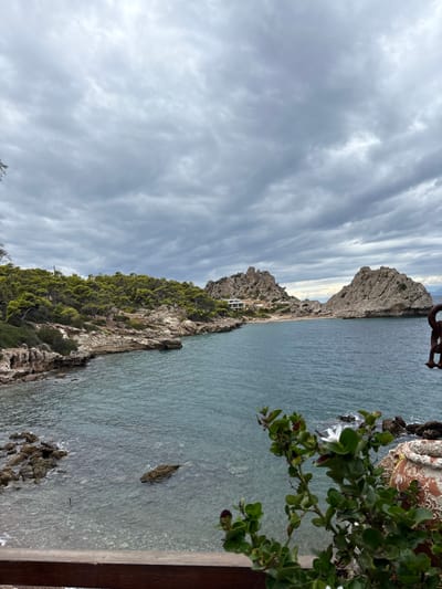 The image depicts a panoramic coastal landscape in the Municipal Unit of Loutraki - Perachora, Greece, under an overcast sky. The top third of the frame is dominated by a dense layer of grey, textured clouds. The central and lower portions feature a tranquil body of teal-blue water, likely a bay or cove, with visible ripples and submerged rocks along the shallow edges.
To the left, a rugged coastline extends from the mid-foreground into the distance, densely covered with dark green trees and scrubby vegetation. Rocky outcrops are visible along the shoreline. In the mid-ground of this coastline, a modern, light-colored building with a flat roof is situated on a slight rise overlooking a small, light-colored sandy beach nestled between rocky sections. Further into the bay, two prominent, jagged grey rock formations rise from the water, defining the outer edge of the cove. Beyond these, in the far distance on the right, faint landmasses are visible beneath the cloudy horizon.
In the immediate foreground, a dark brown wooden railing extends horizontally across the bottom of the image, suggesting a viewpoint from an elevated terrace or balcony. Partially visible on the right foreground are a terracotta-colored pot with an ornate, raised pattern and lush green foliage with small white flowers growing from it, partially obscuring the view of the water. A dark, rusty-colored metal object, possibly a decorative piece resembling an anchor or a chain link, is also partially visible in the upper right corner of the foreground foliage. No individuals or specific actions are depicted in the scene.