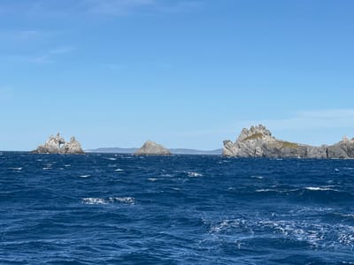 The image shows a body of water with several rock formations jutting out. The water is a deep blue with whitecaps, indicating a choppy sea. The sky is clear and blue. In the background, land is visible. The rock formations vary in size and shape, with one on the left having a natural archway. The scene appears to be a coastal landscape.
