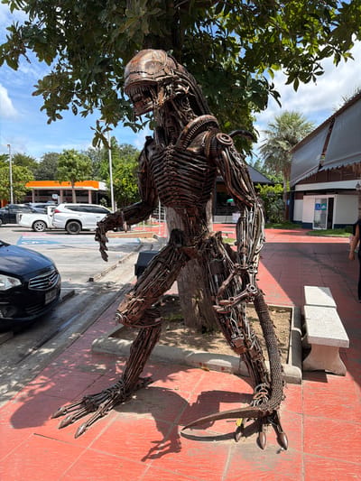 An outdoor shot captures a large, detailed sculpture of an alien from the movie "Alien." The sculpture is made of metal and is situated on a red-tiled pathway in front of a building with a gray, sloped roof and outdoor seating areas. The sculpture stands under the dappled shade of a large tree with green leaves against a bright blue sky with scattered white clouds.

In the background, there are parked cars, including a white SUV and a black sedan. The buildings in the background appear to be commercial establishments, possibly restaurants or shops, with awnings. To the right, a woman wearing a black shirt and black pants is partially visible, walking away from the camera. To the left of the sculpture, there is a parking lot with painted lines. The overall scene suggests a public space in a warm climate, consistent with Tha Ruea, Thailand.