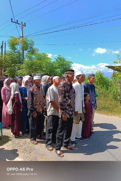 In Blangkutacoh, Indonesia, a group of people, predominantly men wearing traditional attire and white peci caps, stand in a line outdoors on a sunny day. The men are dressed in various patterned shirts and trousers. Some women in the background wear hijabs in shades of pink and blue. A young girl in a burgundy dress is also visible among the group. The environment includes a road with a dirt shoulder, sparse vegetation, trees, and utility poles with electrical wires overhead, under a clear blue sky with scattered clouds. The scene appears to be a gathering, possibly for an event or ceremony.