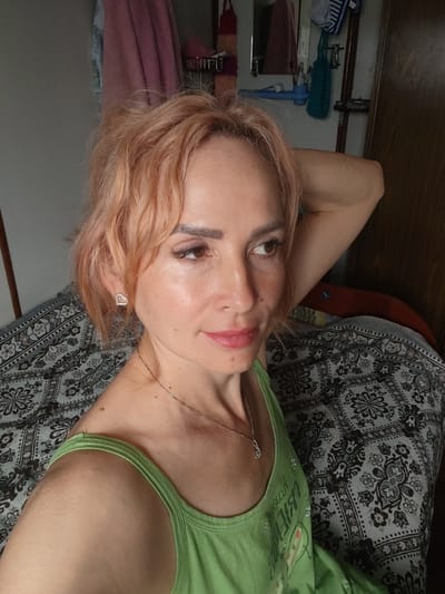 The image shows a close-up, waist-up view of a woman with light-colored hair, wearing a green tank top and a silver necklace. She has a heart-shaped earring in her left ear. Her right arm is bent and resting behind her head. The woman is looking off to the right side of the frame, with her head tilted slightly. The background appears to be an indoor setting, with a bed with a patterned comforter visible. A closet door and some items hanging on the wall are also present. There is no indication of the location being in Shalsky, Russia, or any specific city.