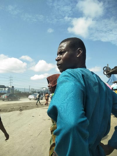 The image depicts an outdoor scene in Ajah, Nigeria, on a sunny day with a clear blue sky and scattered white cumulus clouds. In the foreground, positioned to the right of center, an adult male with dark skin and short, dark hair is shown in right profile, looking forward. He wears a long-sleeved teal garment. His face and forehead appear glistening, possibly from sweat. Directly behind him and slightly to his left, a second individual, also with dark skin, is partially visible. This person wears a brown head covering and a garment with a yellow and green patterned fabric.
The middle ground, to the left of the primary subjects, consists of a dusty, unpaved stretch of ground. Further back, a paved road is visible with multiple vehicles, including a dark-colored sedan and a silver sedan, indicating traffic. Several pedestrians are discernible on or near the paved road, including one person in a light-colored outfit carrying a backpack. In the far background, industrial or modern buildings are visible, along with large utility poles and overhead power lines. To the right edge of the frame, behind the main subjects, a blue tarp structure, possibly a market stall or shelter, is partially visible, with a metallic-looking object featuring a series of connected circles extending upwards from it.