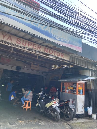 The image shows a motorcycle repair shop in Kecamatan Cimahi Selatan, Indonesia, with a sign above the entrance that reads "Federal Center" and "Super Motor". The shop is located in a narrow alley, and the entrance is obscured by a large number of electrical wires strung overhead. There are three motorcycles parked in front of the shop, and several people are present, including a mechanic working on a motorcycle, and a man sitting on a small orange stool. A small wooden coffee stand is located to the right of the shop. The image was likely captured during the day, as the lighting conditions appear bright.
