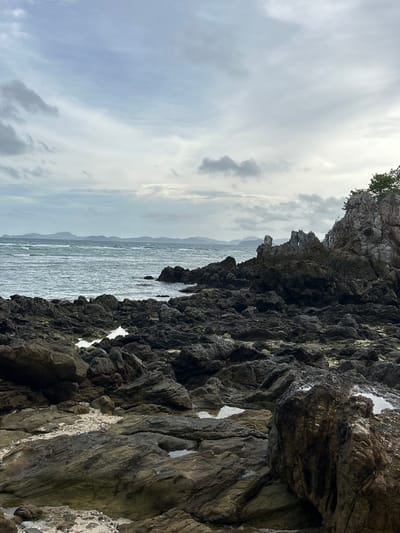 The image depicts a rocky shoreline under a cloudy sky. The foreground is dominated by dark, jagged rocks of various sizes, some wet and reflecting small pools of water. Patches of sand and smaller pebbles are visible between the larger rocks. In the mid-ground, the sea stretches out, with gentle waves lapping against the rocks. The water is a muted blue-green color, and ripples are visible on its surface. In the distance, a series of low-lying islands or hills are silhouetted against the horizon. The sky is overcast with layers of grey and white clouds, creating a diffused light over the scene. To the right of the image, a larger rock formation rises, with sparse vegetation on its summit. The overall impression is one of a rugged, natural coastal landscape in Tambon Phru Nai, Thailand, likely captured on an overcast day. There are no people or visible human activity in the image.