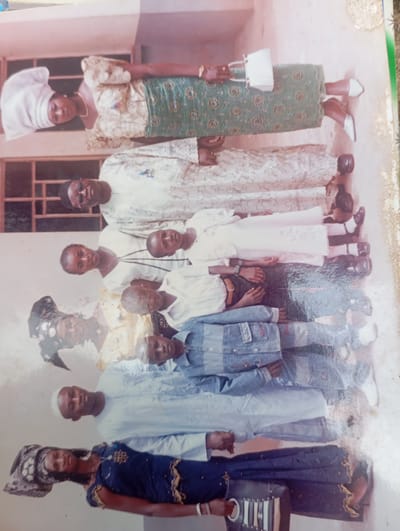 The photograph, taken in Karsana 2, Nigeria, depicts a group of people, appearing to be a family, posed for a formal portrait. The group is arranged in rows, with some individuals standing and others seated or partially visible in the background.
In the front row, a woman in a dark blue dress with intricate gold detailing and a patterned headscarf is holding a dark handbag with white accents. To her right and slightly behind her, a man in a white tunic and cap looks towards the camera.
The middle rows feature several individuals, including two boys dressed in denim jackets and shirts. One boy is wearing a watch on his left wrist. Another individual, possibly an older child or young adult, is wearing a white shirt. Further back, a woman in a white headscarf and ornate patterned clothing is visible, holding a white purse. A man in a white kaftan and glasses is also present in this section of the group.
The background suggests an outdoor setting, possibly a courtyard or an area near a building, with a wall or structure partially visible on the left side of the frame. The lighting indicates it is daytime. The overall tone of the photograph is serious and posed, consistent with a formal family gathering or event.