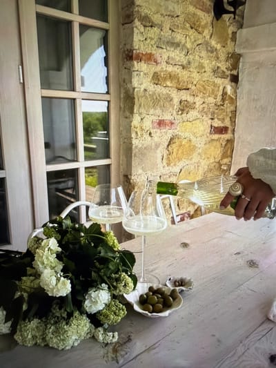 The image captures a moment of beverage pouring in what appears to be a rustic yet elegant setting, located in Ros Na Rí, Ireland. The scene is presented from a slightly elevated, overhead perspective, with the original image rotated ninety degrees counter-clockwise.

In the center of the frame, a person's hand is visible, clad in a white or off-white long-sleeved garment, holding a clear glass bottle. A light-colored, possibly beaded, ring adorns the middle finger of the hand. The bottle, containing a light-colored liquid (likely white wine), has a green foil or label towards its base, and is tilted to pour into one of two empty clear wine glasses below. These glasses are adorned with translucent white ribbons tied around their stems.

To the right, on a light-colored, possibly painted wooden surface, sits a small, white, organic-shaped bowl filled with green olives. Another smaller, similar bowl with a few olives is next to it. In the foreground, a substantial arrangement of white and light green hydrangeas or similar cluster flowers, mixed with dark green foliage, is displayed in a partially visible white vase or container.

The background features a rustic stone wall composed of irregular yellowish stones and scattered reddish bricks. To the lower left, a large multi-paned window or glass door with light-colored wooden frames reflects greenery from outside, indicating daylight conditions. A dark, metallic, wall-mounted object is partially visible on the stone wall to the far left.

The overall atmosphere suggested by the elegant flowers, wine, and olives, combined with the rustic architecture, is one of a relaxed social gathering or preparation for such an event. No visible text is present in the image.