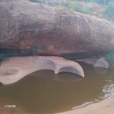 A large, dark-colored rock overhangs a pool of murky water. The water flows around sandy formations with sculpted edges, suggesting erosion. Sparse vegetation grows on the rock's edge and along the water bank. The scene conveys a sense of a natural, weathered landscape, perhaps a riverbank or stream. A "TECNO" watermark is visible in the lower left corner.