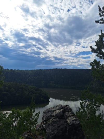 The image depicts a scenic view from a rocky outcrop overlooking a lake and a dense forest. The sky is filled with a dramatic mix of white and gray clouds, with patches of blue sky visible. In the foreground, a large, dark gray rock formation dominates the lower right quadrant, with some green vegetation growing around its base.
Beyond the rock, a tranquil body of water is visible, reflecting the cloudy sky. The lake is surrounded by a thick, dark green forest that stretches across the horizon. The trees are densely packed, creating a solid wall of foliage. The overall impression is one of natural beauty and solitude.
This landscape is located in the vicinity of Sim, Russia.