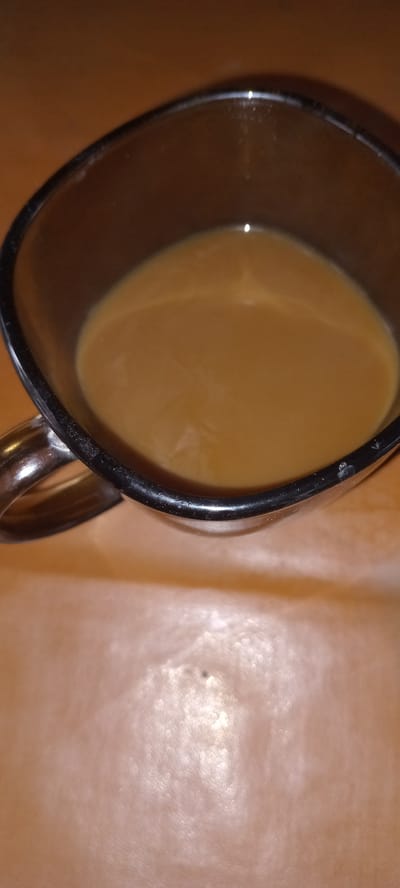 The image shows a close-up, high-angle view of a glass mug filled with a light brown liquid, likely coffee with milk or cream, resting on a wooden surface. The mug has a dark rim and a metallic handle. The background is a blurred, warm-toned surface, possibly a table or countertop. There are no people present in the image. The scene is indoors. The image does not contain any information to determine its location in the city of Koviahy, Ukraine, nor does it provide any details related to forensic analysis or an event.