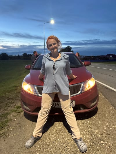 A woman with curly red hair stands in front of a red car on the side of a road at dusk. She is wearing a light blue button-down shirt and beige pants, with dark clogs on her feet. Her hands are resting on the hood of the car. In the background, two other individuals are visible near the car, one standing and looking away, and another peeking from the passenger side window. The environment is rural, with a grassy field to the left and houses with dark roofs visible in the distance to the right. A streetlamp is illuminated near the center of the image. The car's headlights are on. The license plate of the car, partially visible, has Russian characters and numbers including "9". The location is the city of Nikolskoye, Russia.