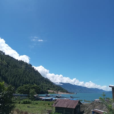 The image shows a picturesque landscape of a lake in Mendale, Indonesia, on a bright, sunny day. The sky is a vibrant blue with fluffy white clouds scattered across it. In the foreground, there is a small wooden structure with a rusted corrugated metal roof. To the left of this structure, a lush green hillside covered in dense trees slopes down towards the water.
The lake itself is a beautiful turquoise color, with calm waters reflecting the blue sky. Along the shoreline, there are several small houses and wooden structures built on stilts, some with colorful roofs. These appear to be part of a small village or settlement on the water. In the distance, more mountains can be seen rising beyond the lake, their peaks partially obscured by clouds. The overall scene conveys a sense of tranquility and natural beauty.