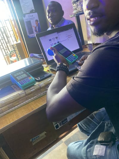 The image shows two people in what appears to be a bar or restaurant setting. One person, whose face is partially visible, is wearing a dark shirt and is looking towards a tablet. They are holding a green payment terminal. The other person is wearing a denim shirt and has a Louis Vuitton key pouch attached to their shirt. A handwritten note with "Anty Sum 37,000" is also visible on their shirt.
The scene is lit with artificial light, creating a dim atmosphere. The tablet displays some charts and data. There are also several other objects on the table, including a phone and what looks like a receipt or printed material with the text "I&a STAY CONNECTED". Another sign visible is "FRAN". The overall impression is that of a transaction or business interaction taking place. The clothing and the presence of a payment terminal suggest a commercial environment. The handwritten note with a monetary value might indicate a sale or a debt payment.