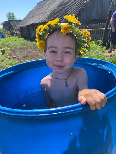 A child is inside a blue plastic barrel, likely filled with water. The child is wearing a crown of dandelions and a necklace with a cross. The barrel is located outdoors near a grassy area and a wooden building. The location appears to be in a rural or semi-rural environment. It is daytime, and the lighting indicates sunny conditions. Based on the prompt, the location is in the city of Узяр, Russia.