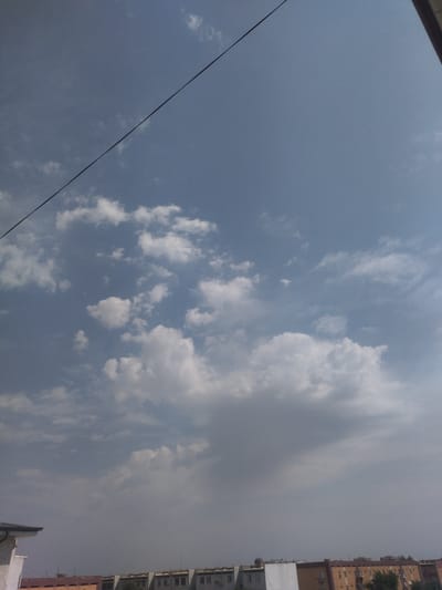 The image shows a view of the sky and buildings in the city of Bekabad, Uzbekistan. The sky is blue with scattered white clouds. A dark power line runs diagonally across the top of the image. Below the sky are several buildings, appearing to be apartment complexes or commercial structures, with visible windows and roofs. Some trees are also visible in the foreground near the buildings. The overall scene depicts an urban environment on a cloudy day.
