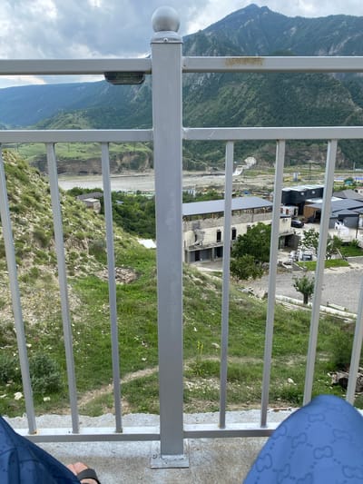 The image shows a view from a balcony or elevated walkway overlooking a mountainous and partially developed landscape, identified as Chalda, Russia. In the foreground, a metal railing, painted gray and showing some signs of rust, divides the view. Behind the railing, the terrain slopes downwards, covered in sparse green vegetation and rocks. Further in the background, a river or large body of water is visible, with a valley carved into the hills.
Across the valley, there are several buildings in various stages of construction and completion. Some appear to be modern structures with dark facades, while others are more traditional brick buildings. Construction equipment, including what looks like a crane and a bulldozer, are visible on a construction site. Further in the distance, more buildings and some signs of infrastructure, like power lines, can be seen. The entire scene is set against a backdrop of large, tree-covered mountains under a cloudy sky.
In the extreme foreground, the bottom edge of the image shows a person's legs wearing blue pants with a repeating pattern, and a sandal on one foot, indicating that the photo was taken by someone sitting or standing on this elevated viewpoint.