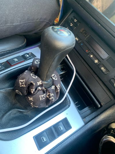 A close-up, eye-level, medium shot captures the interior of a car, focusing on the gear shift and part of the central console. The gear shift lever is black and topped with a round knob displaying gears, with a white cord snaking around its base. The base of the gear shift is adorned with a black scrunchie featuring a repeated white pattern of interlocking "LV" monograms.

To the right of the gear shift is a portion of the car's dashboard, including several black buttons with symbols representing climate control functions such as fan speed and air recirculation. One button is labeled "AUTO". Below the climate controls, a dark gray, recessed area, possibly a storage compartment or ashtray, is visible.

To the left of the gear shift, a row of black buttons is integrated into a silver-toned panel. One button shows a red indicator, and another features a white icon of a car window.

The surrounding materials are primarily black, textured plastic and leatherette, typical of a car interior. The lighting suggests daylight, casting subtle shadows and highlighting the textures of the surfaces. The overall impression is a utilitarian view of a car's interior, with a decorative, personalized touch on the gear shift. The location is identified as Madan, Bulgaria.