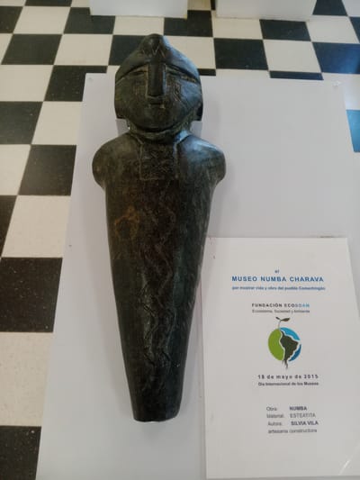 This indoor photograph, rotated 90 degrees clockwise, showcases an exhibit featuring a sculpted object and an informational sign. The central subject is a dark, elongated sculpture, resembling a stylized human or totemic figure with a distinct head, broad shoulders, and a body tapering to a point. Its surface is adorned with engraved patterns. The object rests horizontally on a white display surface.
Beside the sculpture, a white informational sign provides details about the exhibit. The text indicates it is from the "MUSEO NUMBA CHARAVA," dedicated to "mostrar vida y obra del pueblo Comechingón" (showing the life and work of the Comechingón people). The exhibition was sponsored by "FUNDACIÓN ECOSOAM" (Ecosystem, Society and Environment) and took place on "18 de mayo de 2015," coinciding with "Día Internacional de los Museos" (International Museum Day). The artwork is titled "NUMBA," crafted from "ESTEATITA" (steatite or soapstone) by author "SILVIA VILA," described as an "artesana constructora" (artisan builder).
The background features a white wall or display panel and a contrasting black and white checkered floor. The scene suggests an indoor museum or exhibition setting in Villa Carlos Paz, Argentina.