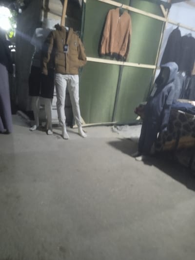 This image captures a market stall or outdoor retail area in the city of Al-Mawasi, Palestinian Territories, under artificial lighting. In the left-center, two headless mannequins are present. One mannequin wears a brown puffer jacket with a visible tag and white pants. Behind it, another mannequin is partially visible, wearing a grey top and a black skirt. To the far left, the lower portion of an individual dressed in dark pants is partially visible. In the right-center, a person, dressed in a dark blue hooded garment with a visible tag, stands or leans beside a display table with a patterned surface. The background features a temporary structure composed of wooden beams supporting a green tarp or fabric wall. A light brown jacket hangs on one of the horizontal wooden beams. Additional dark-colored garments are suspended from a rack to the right. The ground is a light-colored concrete or paved surface.