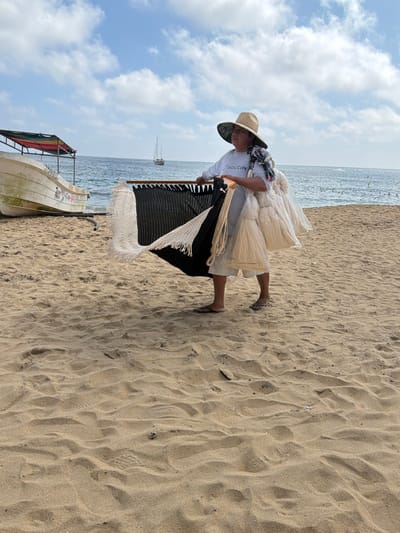 On a sandy beach under a partly cloudy sky, a person wearing a straw hat, printed headscarf, and light-colored clothing carries woven goods. These include cream-colored hammock bags and a black and white blanket with fringe. A small boat is partially visible in the lower left corner. The ocean stretches to the horizon. A sailboat is visible in the distance.