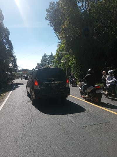 The scene depicts an outdoor road environment in Ka Thaung (Upper), Myanmar, under clear skies with bright sunlight originating from the upper left, causing lens flare. The asphalt road, wet in some areas, extends into the background, featuring a solid white line on its left edge and a solid yellow line delineating the right side.
In the mid-foreground, a dark-colored Toyota Innova minivan, observed from the rear, displays an Indonesian license plate B 1404 SBE and illuminated red tail lights. To the immediate right of the minivan, a white scooter with an orange-rimmed rear wheel is ridden by an individual wearing a black helmet and dark clothing. Further right and slightly behind, a light-colored scooter carries two individuals: the driver wears a white helmet and light shirt, and the passenger wears a brown headscarf and a light-colored top. A red motorcycle with a helmeted rider is visible further back and to the left of the white scooter. Additional vehicles and motorcycles are partially discernible in the distant background.
Both sides of the road are lined with dense green foliage and trees. On the right, streetlights on poles are interspersed among the trees. On the left, a path or shoulder area features indistinct structures and a visible circular white sign with dark lettering. Utility lines stretch horizontally across the upper mid-ground. Strong shadows are cast by the vehicles and trees onto the road surface, indicating the bright, direct sunlight.