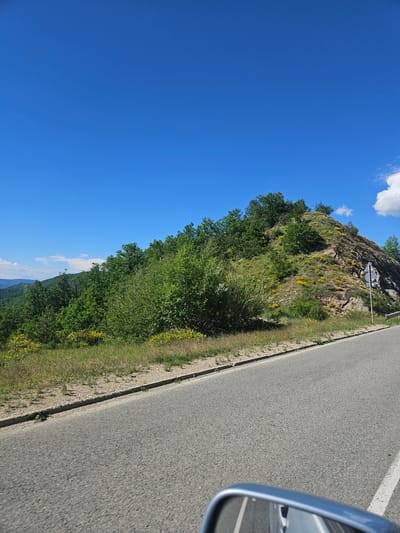 The image depicts a road scene in Liaskovo, Bulgaria. A grey asphalt road occupies the right side of the frame, with a white line marking its edge. A car's side mirror is partially visible on the right. To the left of the road, a grassy verge transitions into a hillside covered in green vegetation, including trees and yellow flowering plants. The top of the hillside reveals rocky terrain with more vegetation. The sky, a clear blue with some white clouds, fills the left side of the image.