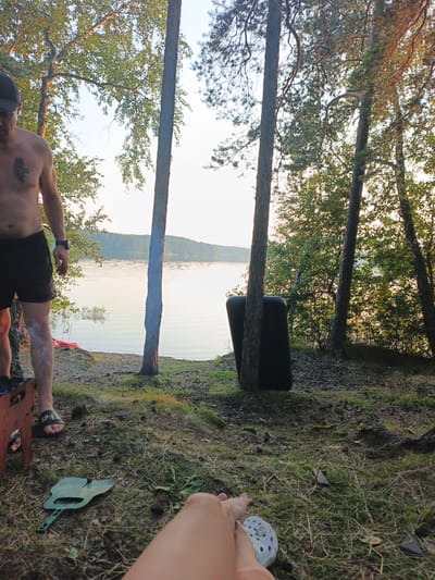 The image depicts an outdoor scene near a body of water, possibly a lake, with a wooded background. The location is identified as Nepryakhinskoye Sel'skoye Poseleniye, Russia.
In the foreground, a man is partially visible, wearing black shorts and a dark baseball cap. He is shirtless, revealing a tattoo on his chest. He is standing near a portable grill, and a white substance appears to be on his lower legs. A flip-flop is on his left foot.
To the right of the man and slightly behind him, two dark, rectangular objects are leaning against trees. These objects resemble large cushions or inflatable mats.
The ground in the foreground is covered with grass, dirt, and fallen leaves. A green shovel-like object is lying on the grass to the left of the frame. In the lower right corner, a person's leg and foot are visible, with a white object resembling a shoe or sandal.
The middle ground features the shoreline of the body of water. The water is calm and reflects the sky and surrounding trees. A faint plume of smoke is visible rising from the area near the grill.
The background consists of a dense forest with numerous trees, many of which appear to be pines and birches. The sky is visible through the tree canopy, with a hazy, bright light suggesting either early morning or late afternoon. The overall atmosphere suggests a campsite or recreational area by the water.