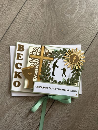 The image displays a close-up, top-down view of a handmade card on a wooden surface. The card features several decorative elements related to a religious ceremony, likely a christening or communion. Prominently featured are large gold letters spelling "BESKO" vertically on the left side of the card. To the right, a decorative gold cross is superimposed on a circular emblem with a smaller gold cross in the center. A silhouette of a man reaching out to a child is depicted in the lower right quadrant of the card's main design. Green foliage elements are scattered around these central motifs. A strip of text in what appears to be Cyrillic script is visible at the bottom of the card, along with a mint green ribbon tied in a bow. The wooden surface suggests a flat lay presentation, possibly for a product or craft display. There is no indication of the city of Belfast or the United Kingdom in the image.