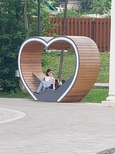 The image depicts a heart-shaped wooden bench in a park-like setting. Two individuals are seated on the bench. One person, wearing a white t-shirt and blue jeans, is sitting upright and appears to be holding something to their face. The second person, wearing a black shirt and blue jeans, is lying down on the bench. The bench is made of curved wooden slats with a white trim along the heart's edge. The background features green trees, a wooden fence, and a paved walkway. A white column is visible on the right side of the frame. The overall environment suggests an outdoor public space. The location is identified as Alikonovka, Russia.
