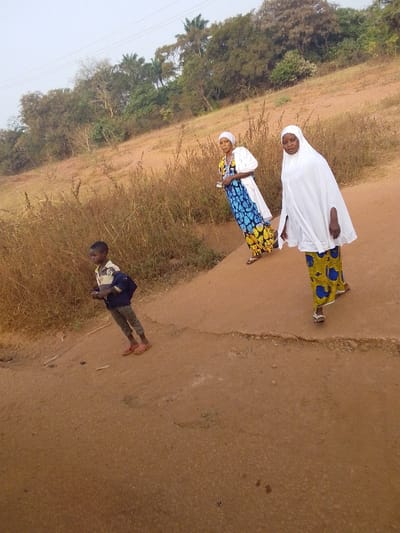 This image depicts three individuals on a reddish-brown dirt path in Oturkpo, Nigeria. A young boy, dressed in a striped shirt, grey trousers, and red sandals, with a blue backpack, stands on the lower left portion of the path. Further up the path, towards the center-left, a woman wearing a white head covering and a long dress with a white bodice and blue, yellow, and black patterned skirt stands, looking towards the left side of the frame. To her right and slightly forward, another woman, clad in a white headscarf and a long white outer garment over a blue and yellow patterned skirt, walks or stands, facing the left. The path is unpaved, with a visible slight elevation difference. An embankment to the left and rear is covered in dry, tall grasses and shrubs, backed by a treeline and a light, hazy sky.